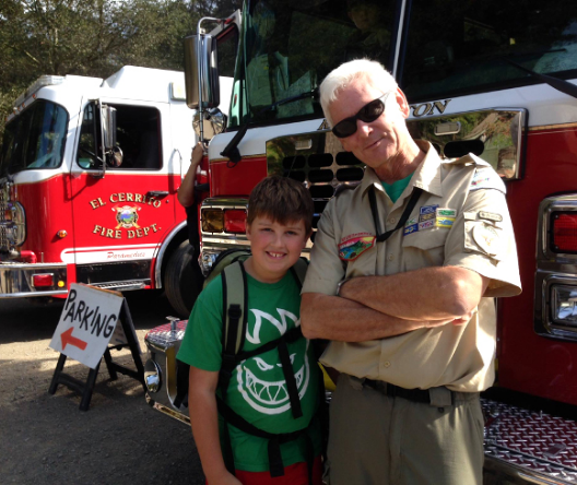 Photo of Jim DeKloe (white hair, sunglasses, scouting uniform) with a young boy in front of firetrucks.
