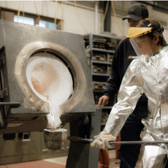 Photo of a student wearing protective gear holding a special cup on a stick while liquid metal is poured into it. An instructor stands close by. Photo credit: IACMI