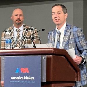 Photo of two men wearing plaid suits standing at a podium with a sign with America Makes' logo on it.