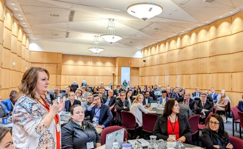 Photo from the 2023 Manufacturing USA Annual Meeting. Woman holding a microphone asking a question with attendees in the background.